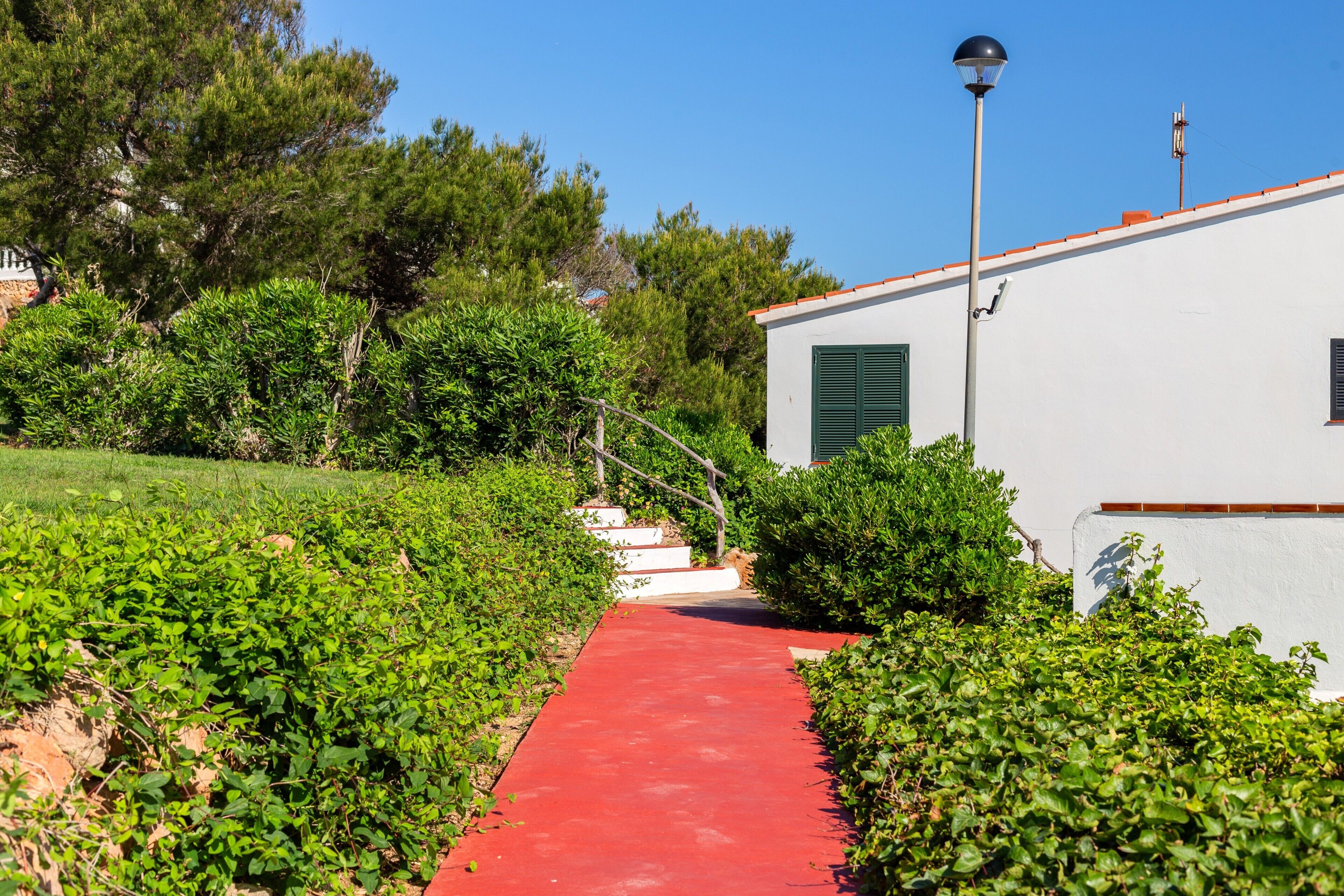 A pathway leading to a white building with green shutters, surrounded by greenery and a clear blue sky.