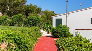 A pathway leading to a white building with green shutters, surrounded by greenery and a clear blue sky.