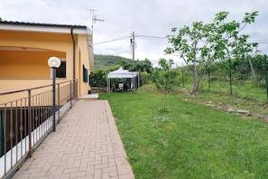 Un camino pavimentado que conduce a un edificio con un balcón, una zona exterior cubierta y un patio con césped y árboles.
