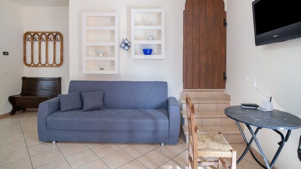 Un salon avec un canapé bleu, un banc en bois, une table à manger, un téléviseur fixé au mur et un escalier menant à une porte en bois.