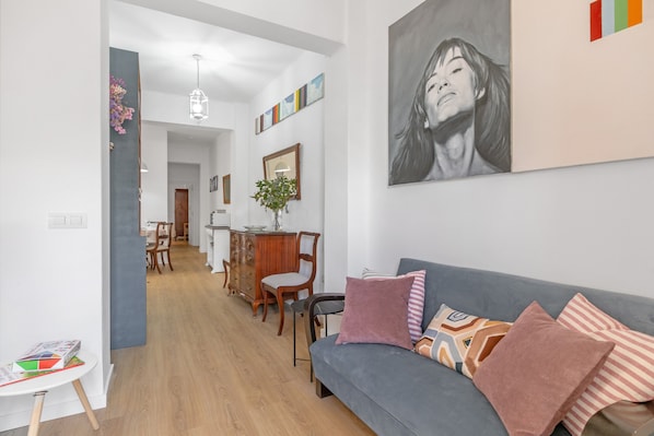 A modern living room with a grey sofa, wooden floor, and a dining area in the background.