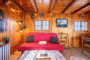 A wooden cabin interior with a red sofa, a bed with a remote control, and a wooden shelf with decorative items.