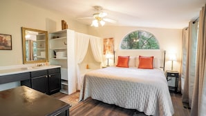 Un dormitorio con cama, tocador, espejo, ventilador de techo y ventana con vista a árboles.