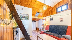 Une pièce chaleureuse aux murs et au plafond en bois, avec un canapé recouvert d’une couverture rouge, une table en bois et une fenêtre donnant sur les montagnes.