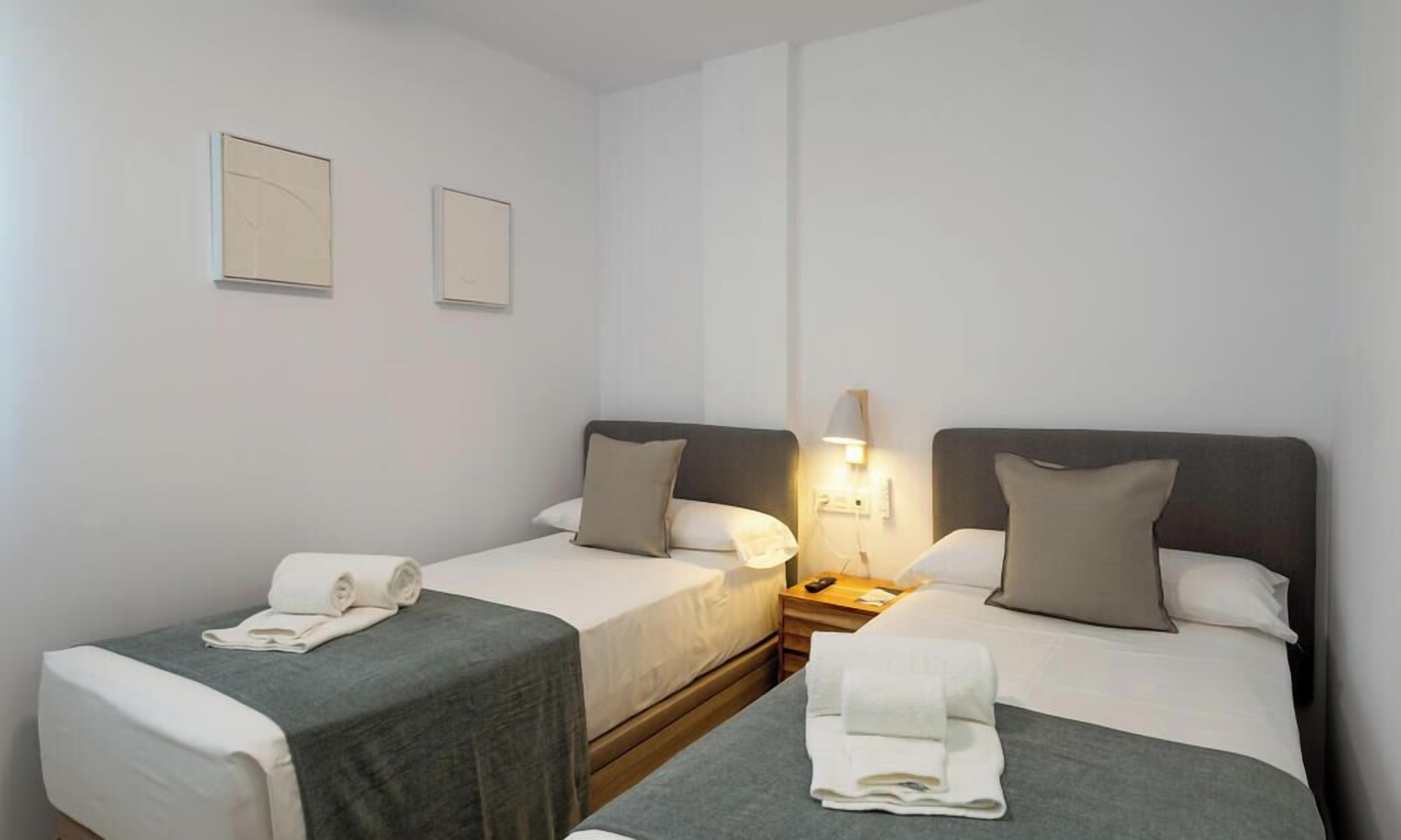 A hotel room with two single beds, each with a grey bedspread and white linens. There is a bedside table with a lamp, a wall-mounted power outlet, and a small nightstand. The room has a clean, minimalist design with white walls and a wooden floor.