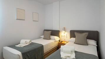 A hotel room with two single beds, each with a grey bedspread and white linens. There is a bedside table with a lamp, a wall-mounted power outlet, and a small nightstand. The room has a clean, minimalist design with white walls and a wooden floor.