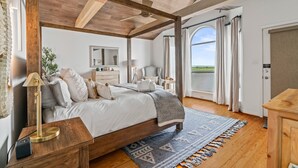 A bedroom with a large bed, wooden ceiling beams, a view of the outdoors, a bedside table with a lamp, and a dresser.
