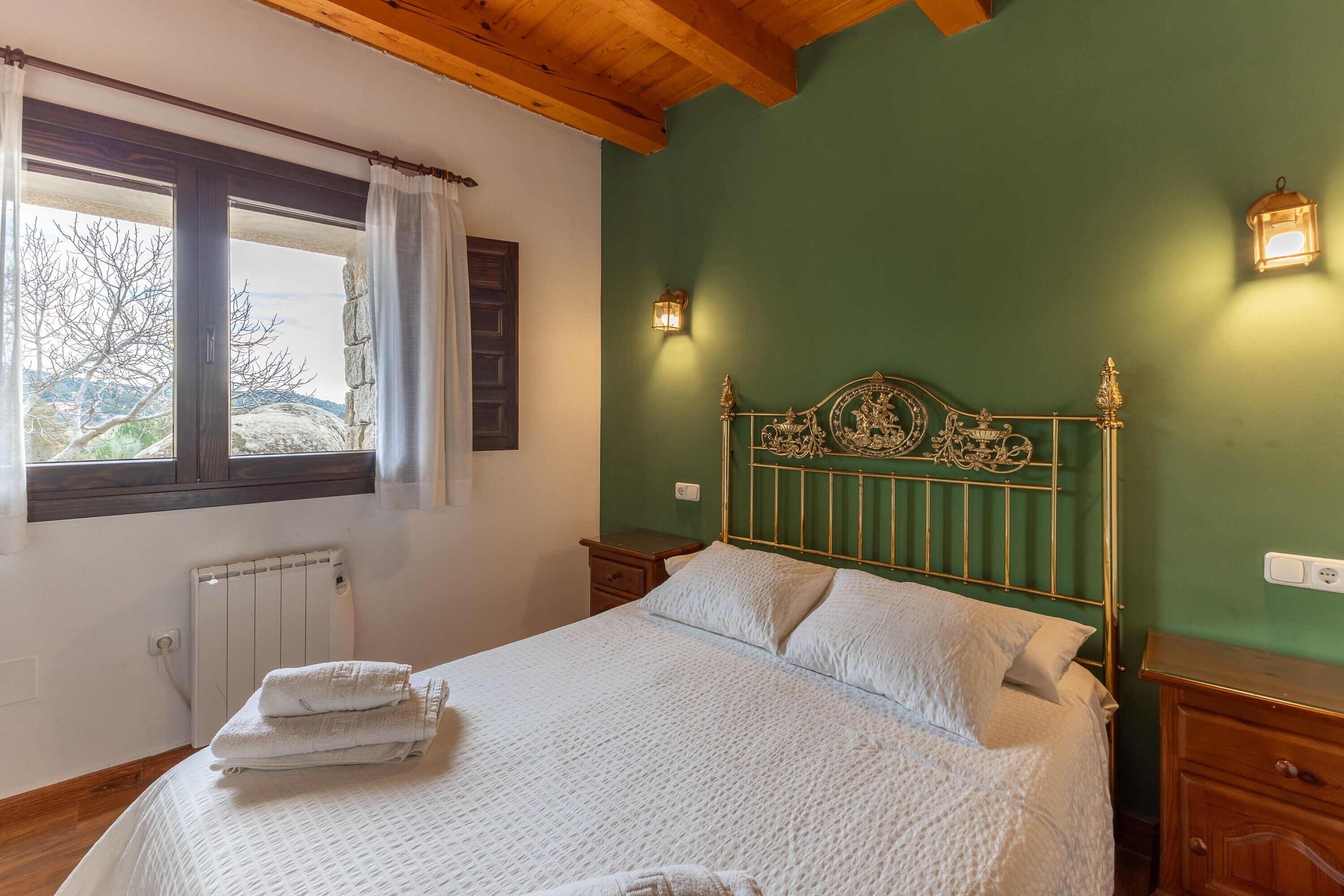 A bedroom with a bed, a window, a radiator, a nightstand, and a wall-mounted light fixture.