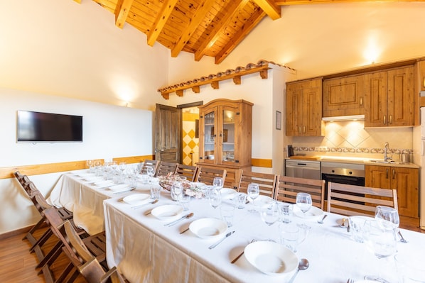 A dining area with a long table set for a meal, wooden chairs, a wooden ceiling, and a kitchen area with wooden cabinets and appliances.