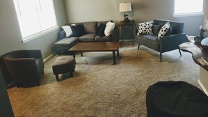A living room with a sectional sofa, a coffee table, and a lamp. There are two windows and an air conditioning unit on the wall. - Serenity's Sanctuary in this quiet, peaceful 2 bdr 1 bath bungalow. Pet friendly (Cushing)