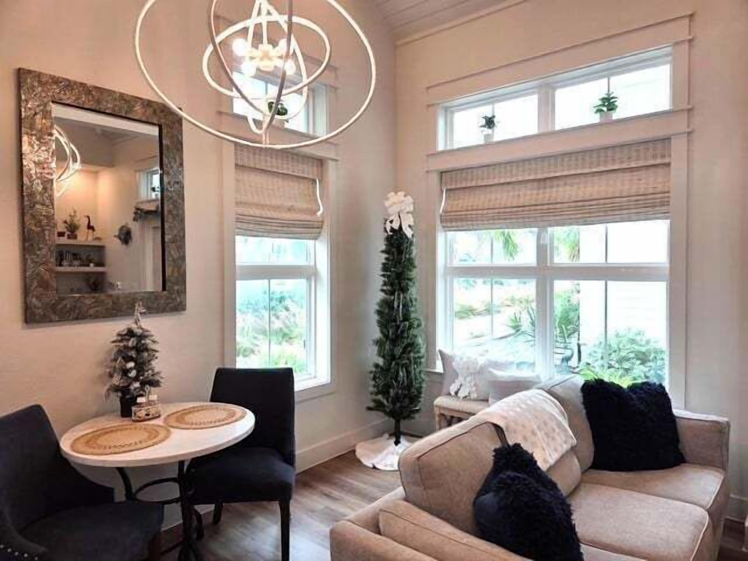 Una sala de estar con una mesa redonda, dos sillas, un sofá con cojines negros, un árbol de Navidad alto y un ventanal con persianas.