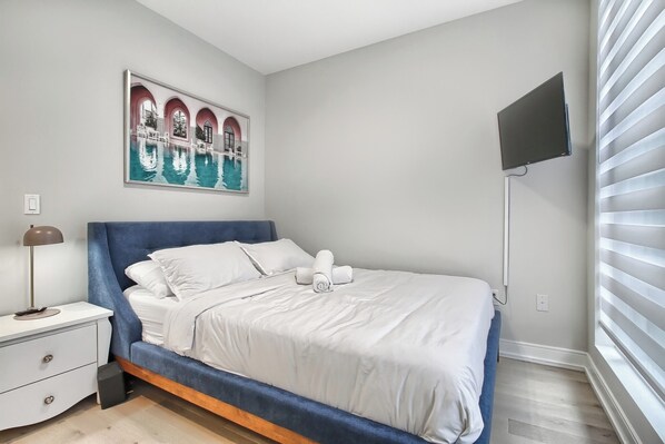 A neatly made bed with white linens, a bedside lamp, and a painting of a pool on the wall.
