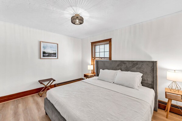 A neatly arranged hotel room with a large bed, a bedside table with a lamp, a framed picture on the wall, and a window with blinds.