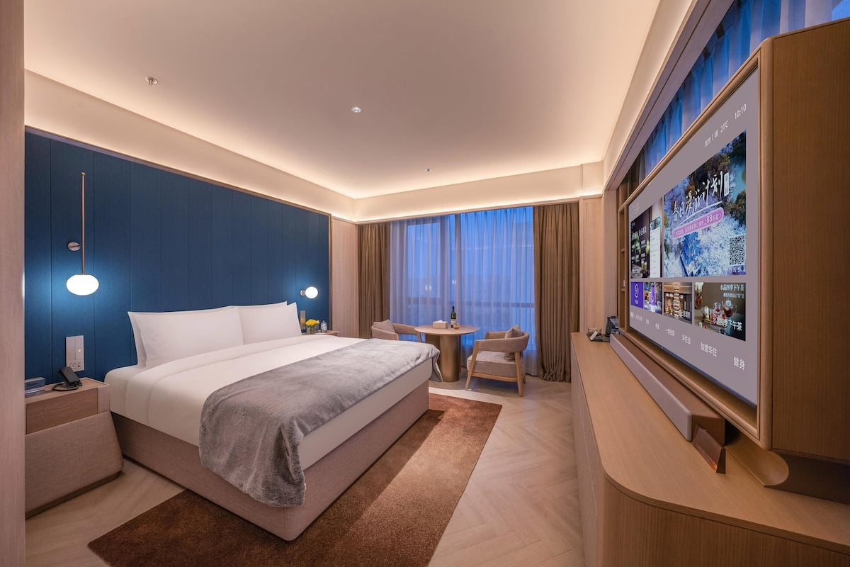 A modern hotel room with a large bed, bedside tables, a TV, and a seating area with a table and chairs.