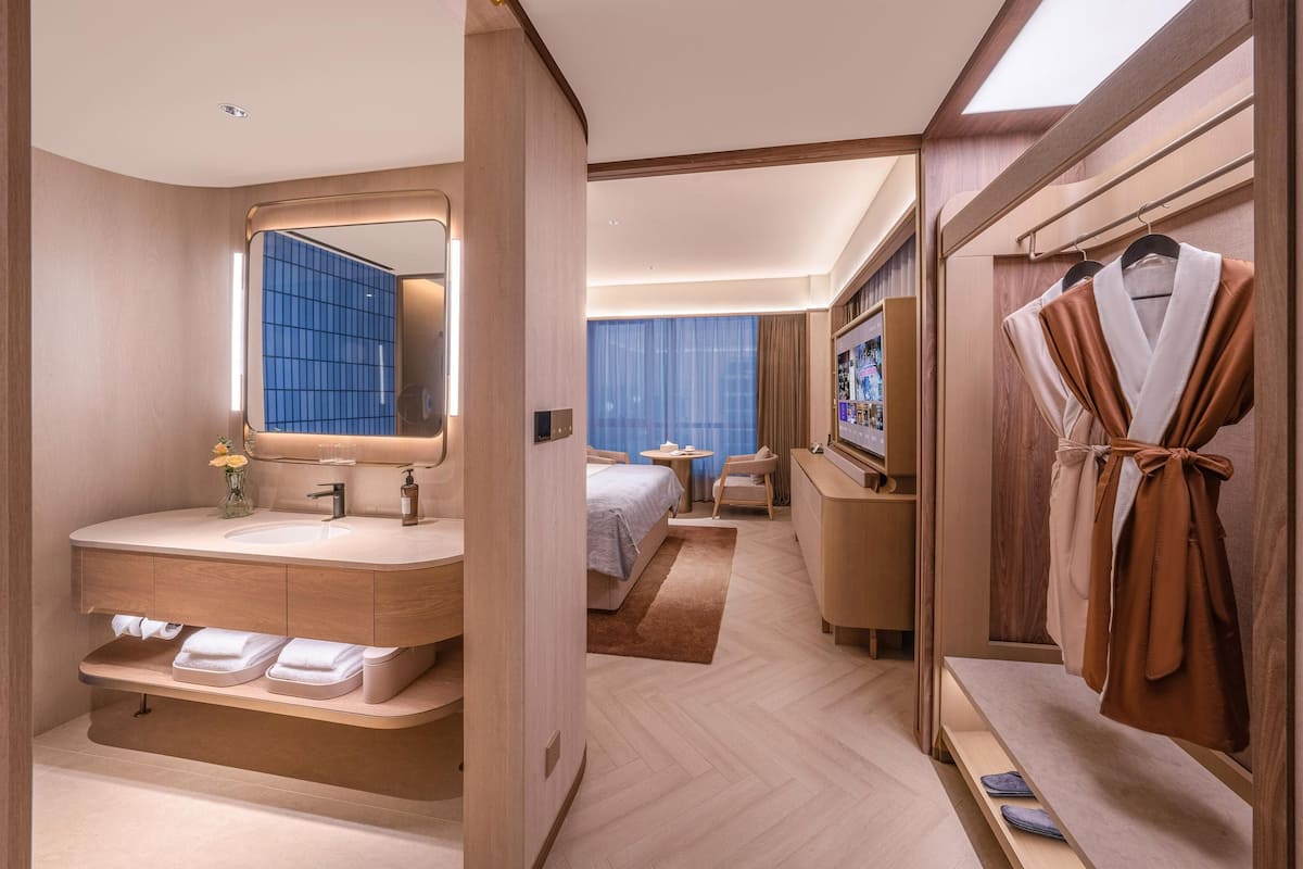 A modern hotel room with a large mirror, a vanity with a sink, a bed with a blue curtain, a TV, and a robe rack.