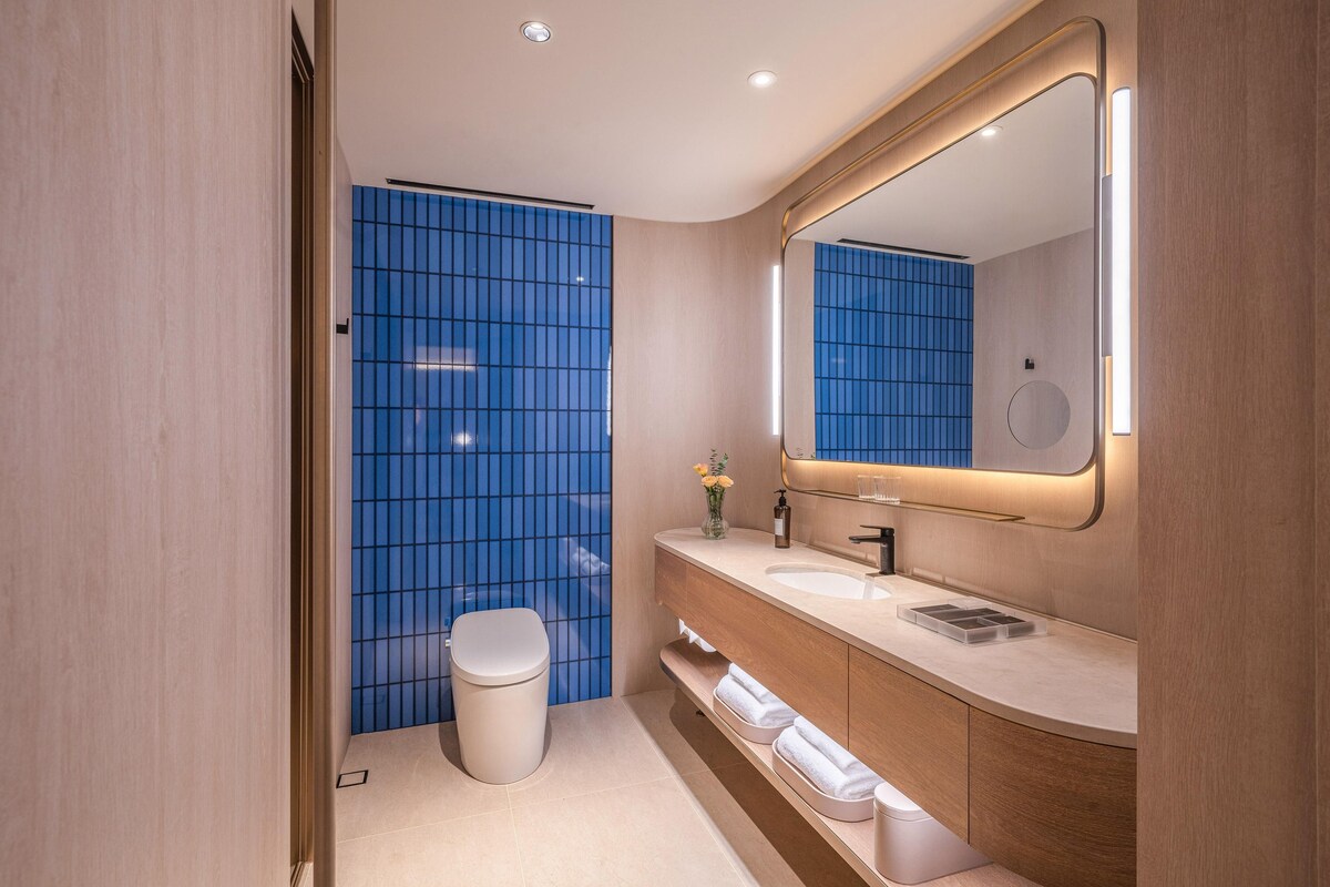 Un baño moderno con pared de ducha en azulejos azules, inodoro blanco, tocador con espejo y lavabo.
