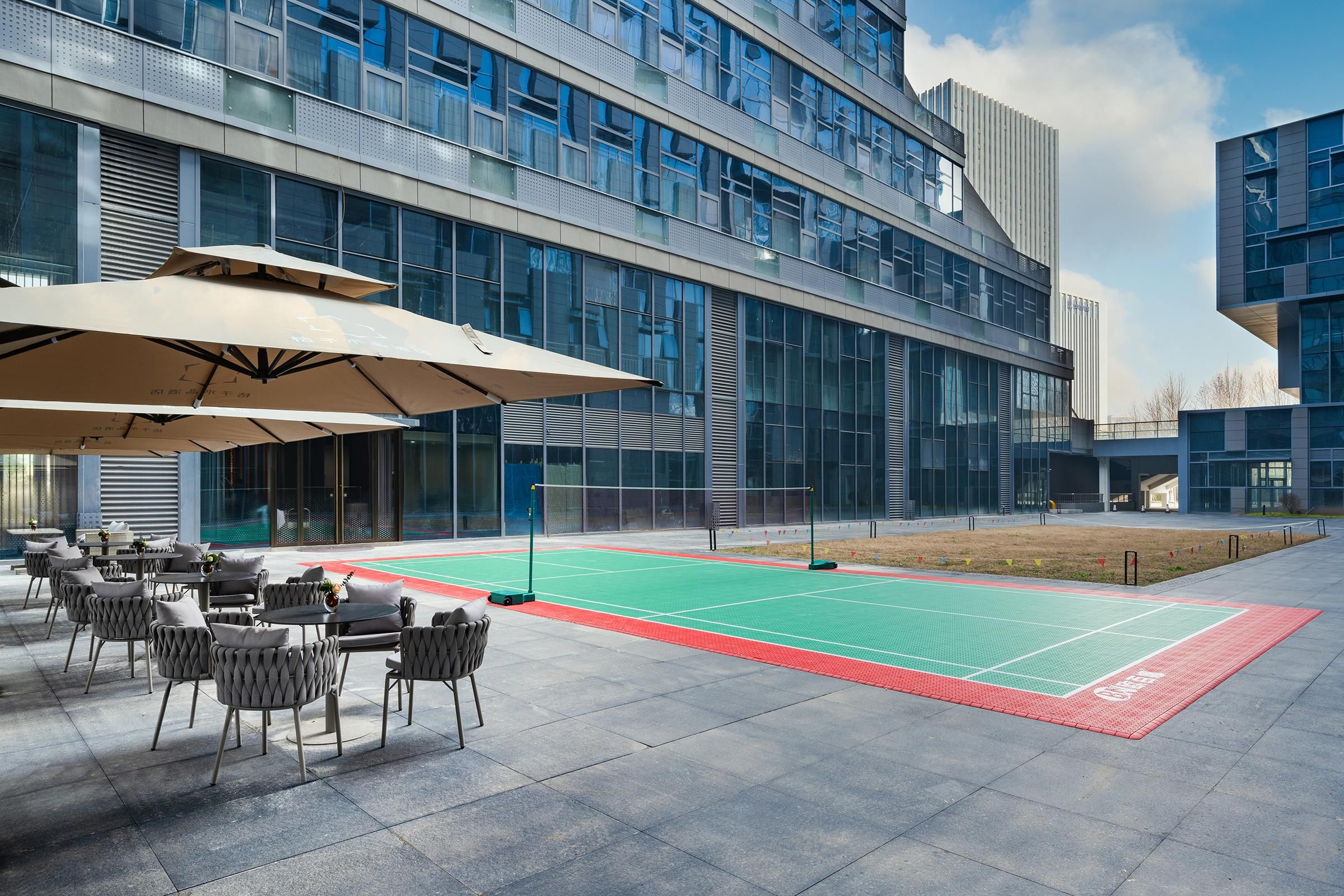 An outdoor seating area with tables and chairs, a tennis court, and a modern building with large windows.