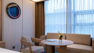 Una habitación de hotel moderna con una mesa redonda, dos sofás, una silla y un ventanal con cortinas traslúcidas.