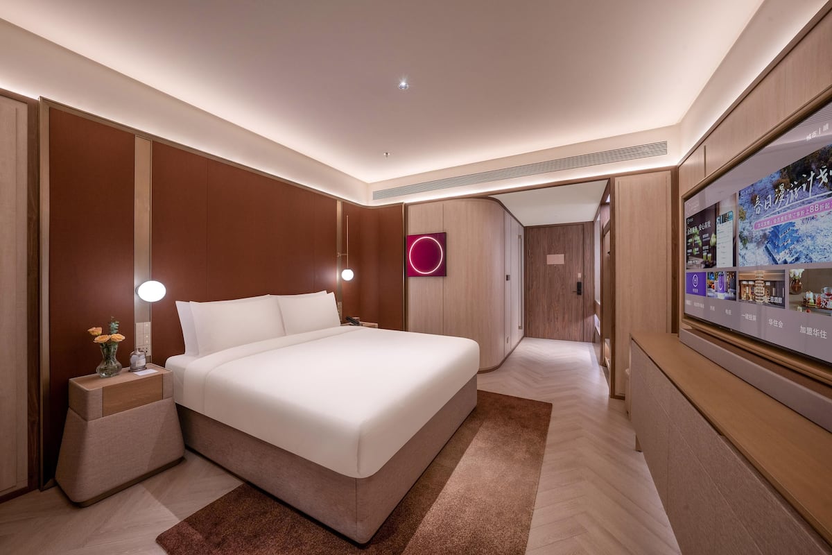 A modern hotel room with a large bed, bedside tables with lamps, a wall-mounted TV, and a wooden wardrobe.