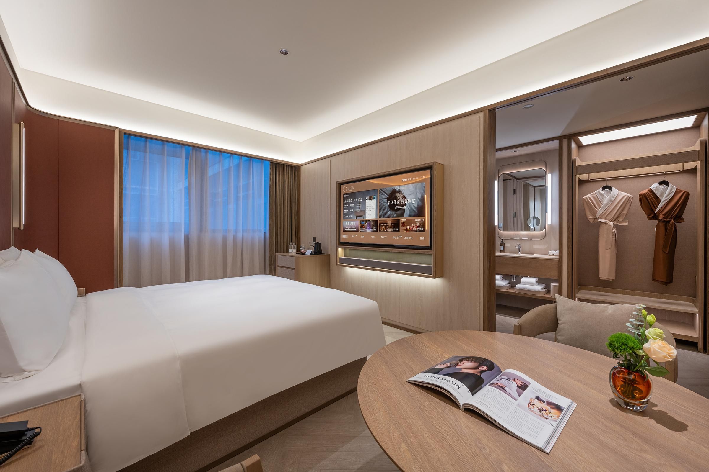 A hotel room with a bed, a desk with a magazine, a mirror, and a TV on the wall.