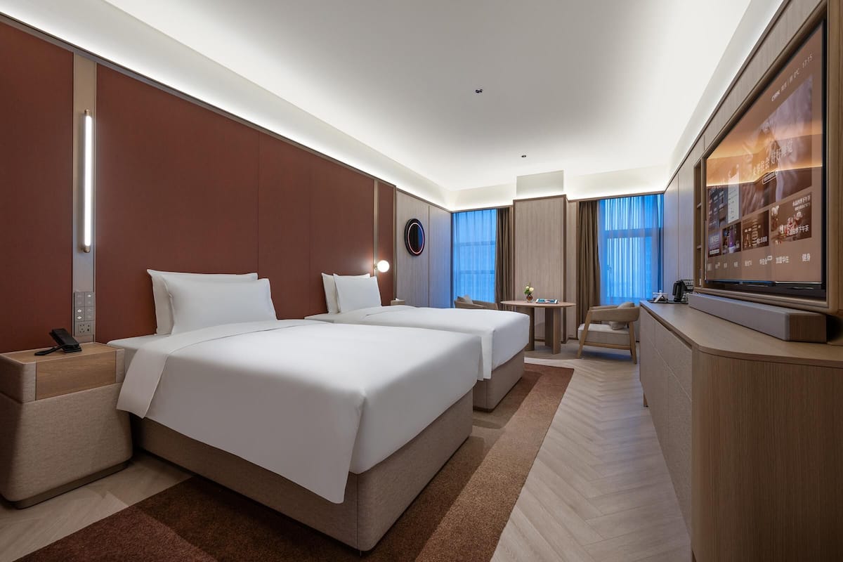 A hotel room with two beds, a desk, a TV, and a large window with curtains.