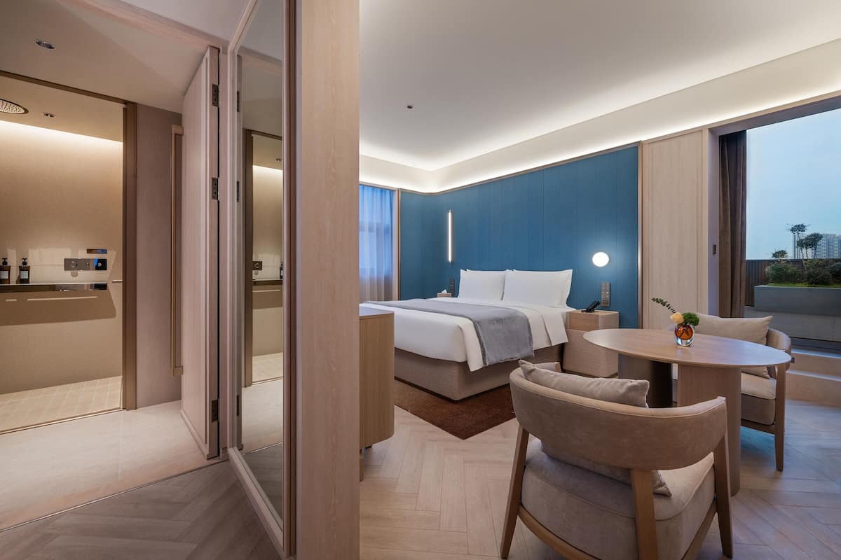 A modern hotel room with a bed, a desk, a chair, and a view of the city.