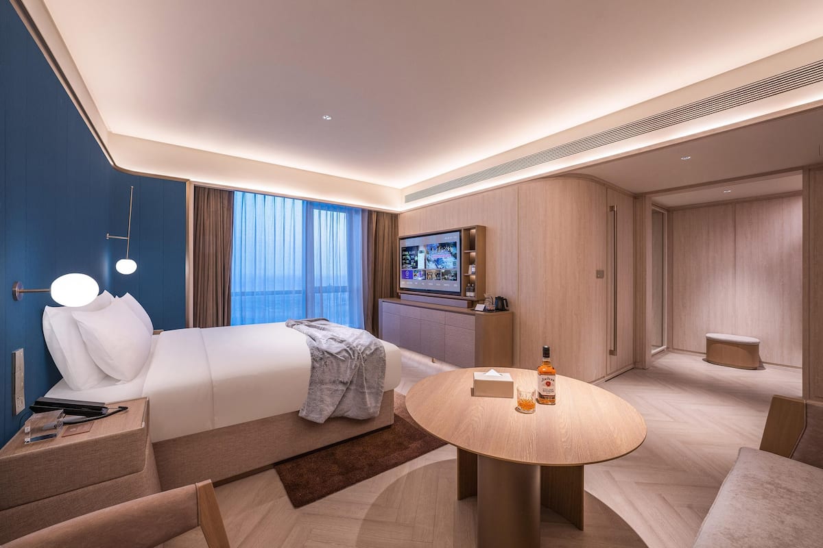 A modern hotel room with a bed, a round dining table, a TV, and a minibar.