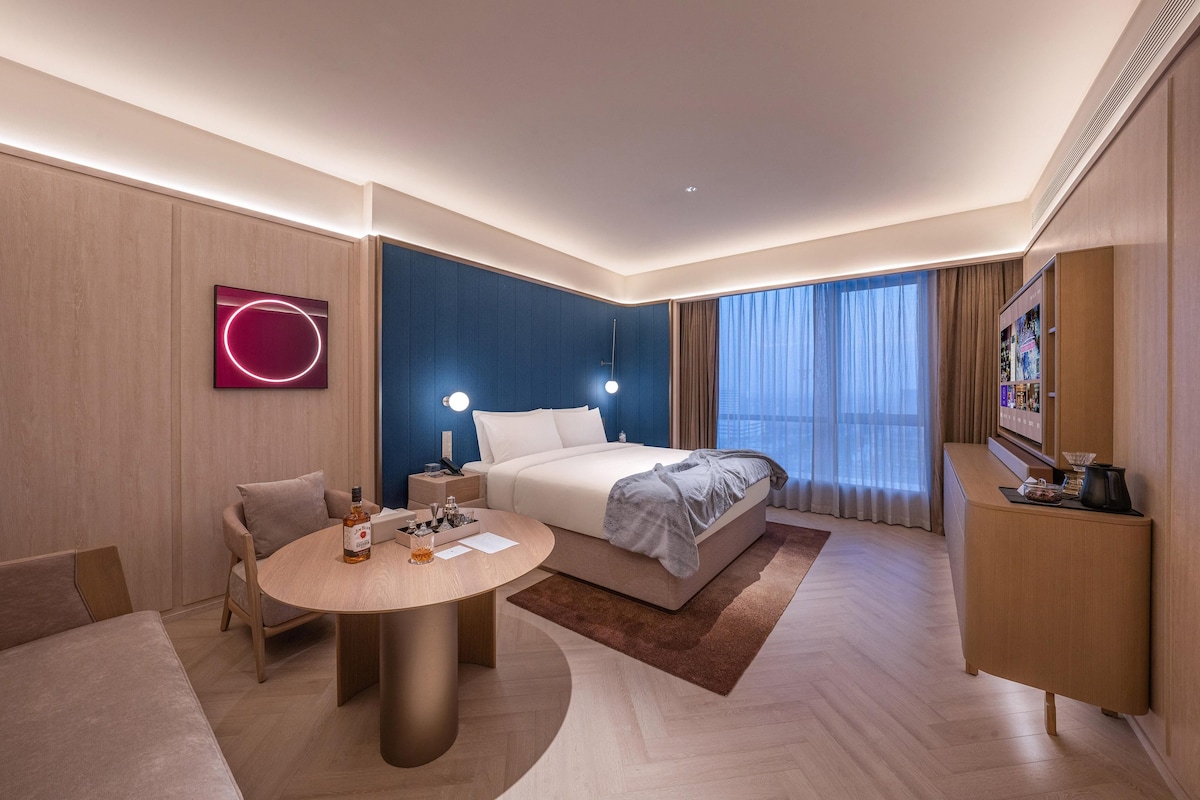 Habitación de hotel con cama, mesitas de noche, escritorio, silla, televisor y ventana con cortinas.