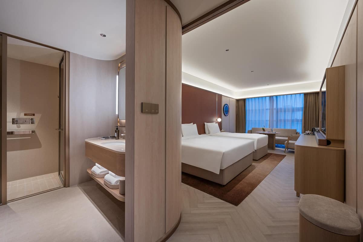 A modern hotel room with a large bed, a separate seating area, a bathroom with a vanity, and a view of the city through large windows.