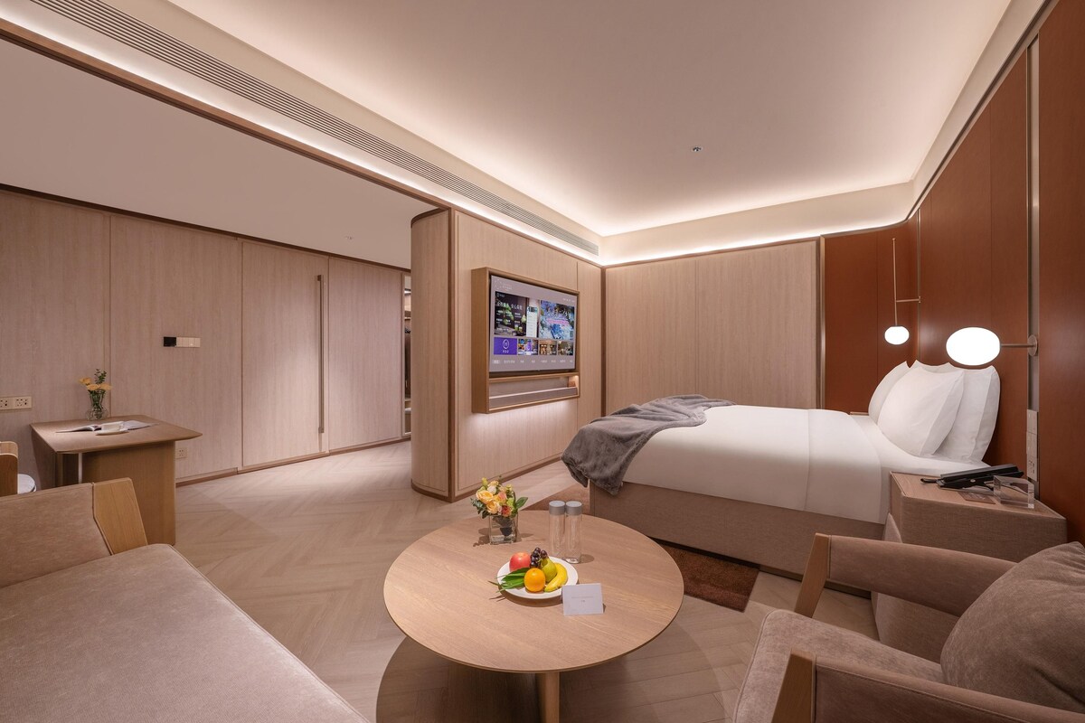 Una habitación de hotel moderna con cama, escritorio, mesa redonda con fruta y televisor en la pared.