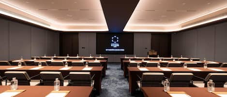 A conference room with multiple tables and chairs, a large screen at the front, and a modern ceiling design.