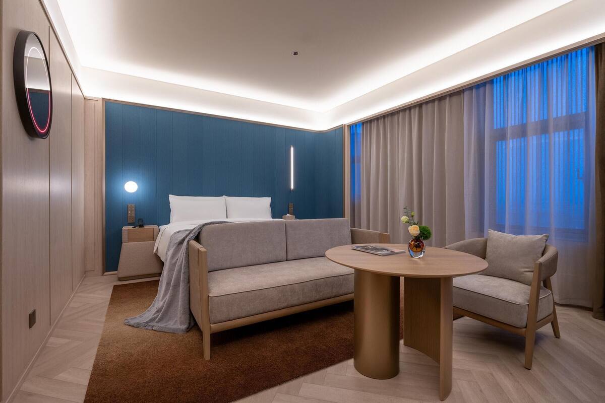 A modern hotel room with a sofa, a round coffee table, and a bed with a nightstand.