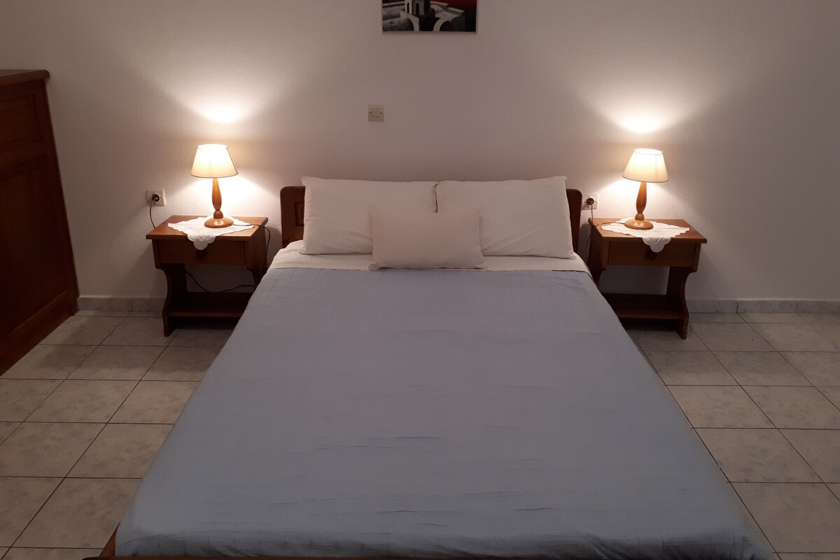 A double bed with white linens, two bedside tables with lamps, a small painting on the wall, and a wooden wardrobe.