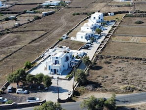 A row of white buildings with a central road, parked cars, and sparse vegetation. - Hotel Paradisos Oia (Santorini)