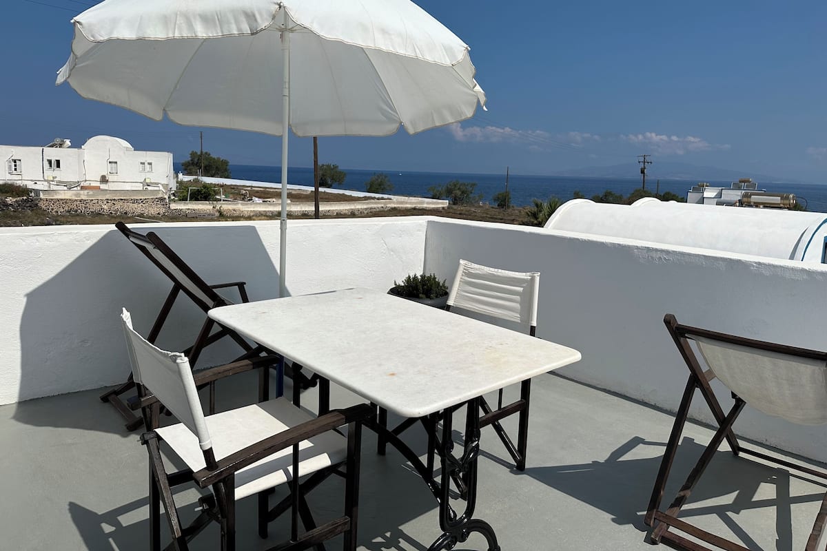 A terrace with a table and chairs, a white umbrella, and a view of the sea and buildings.
