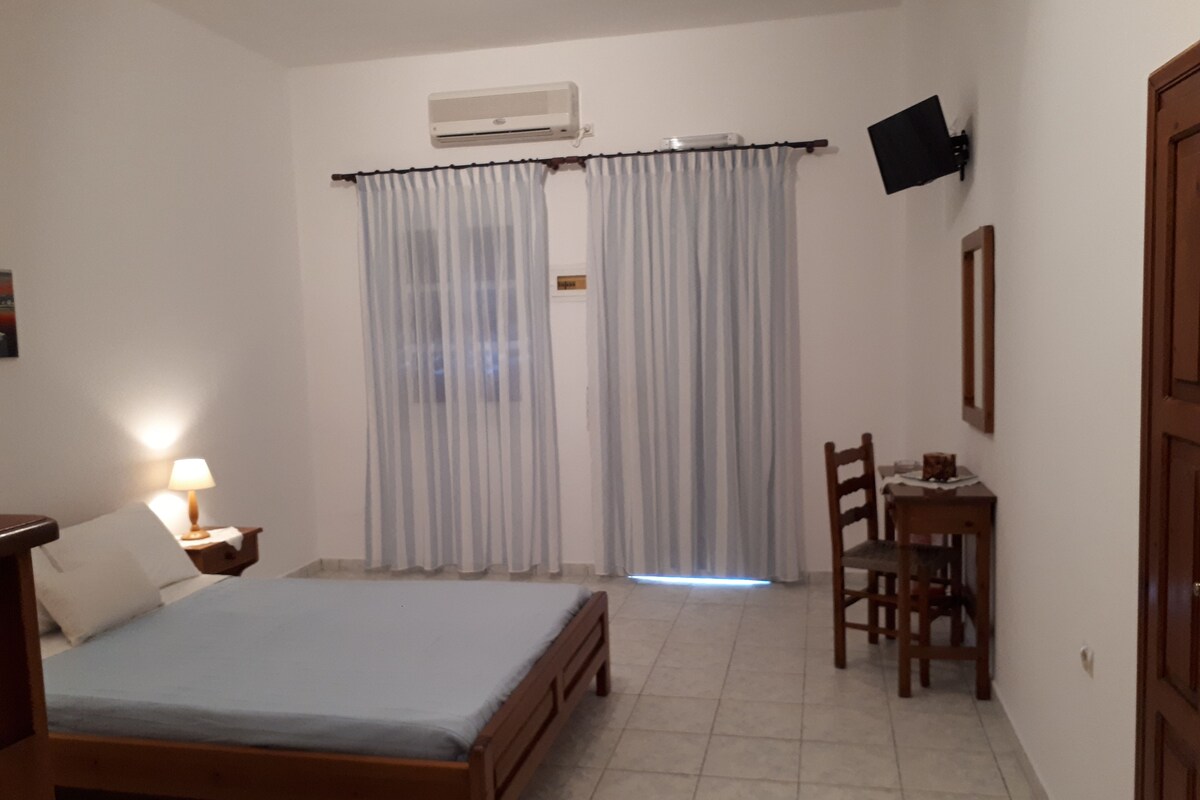 A bedroom with a bed, a nightstand with a lamp, a chair, a desk, and a TV mounted on the wall.