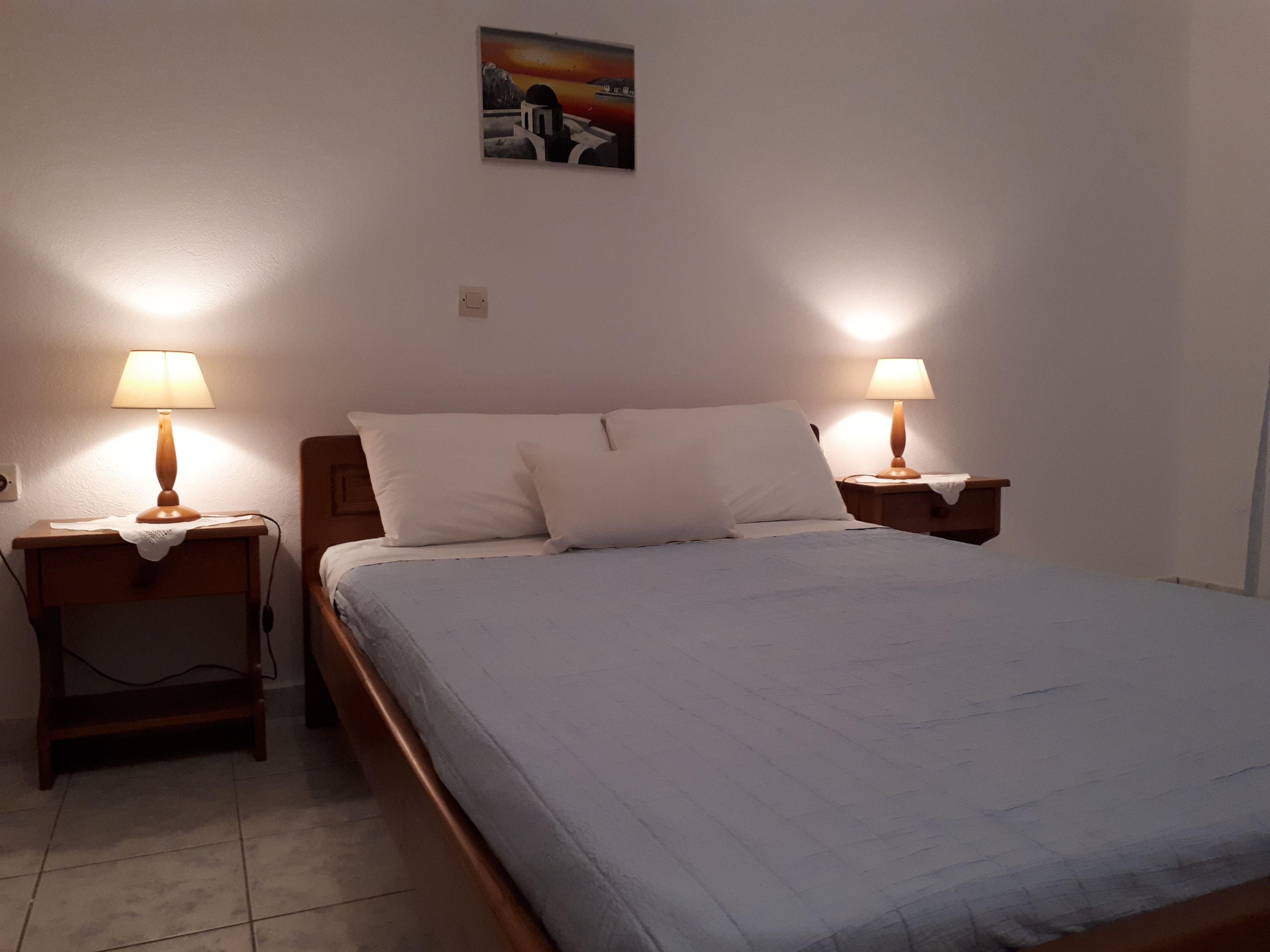 Una cama bien hecha con dos lámparas de noche, una mesita auxiliar pequeña y un cuadro colgado en la pared.