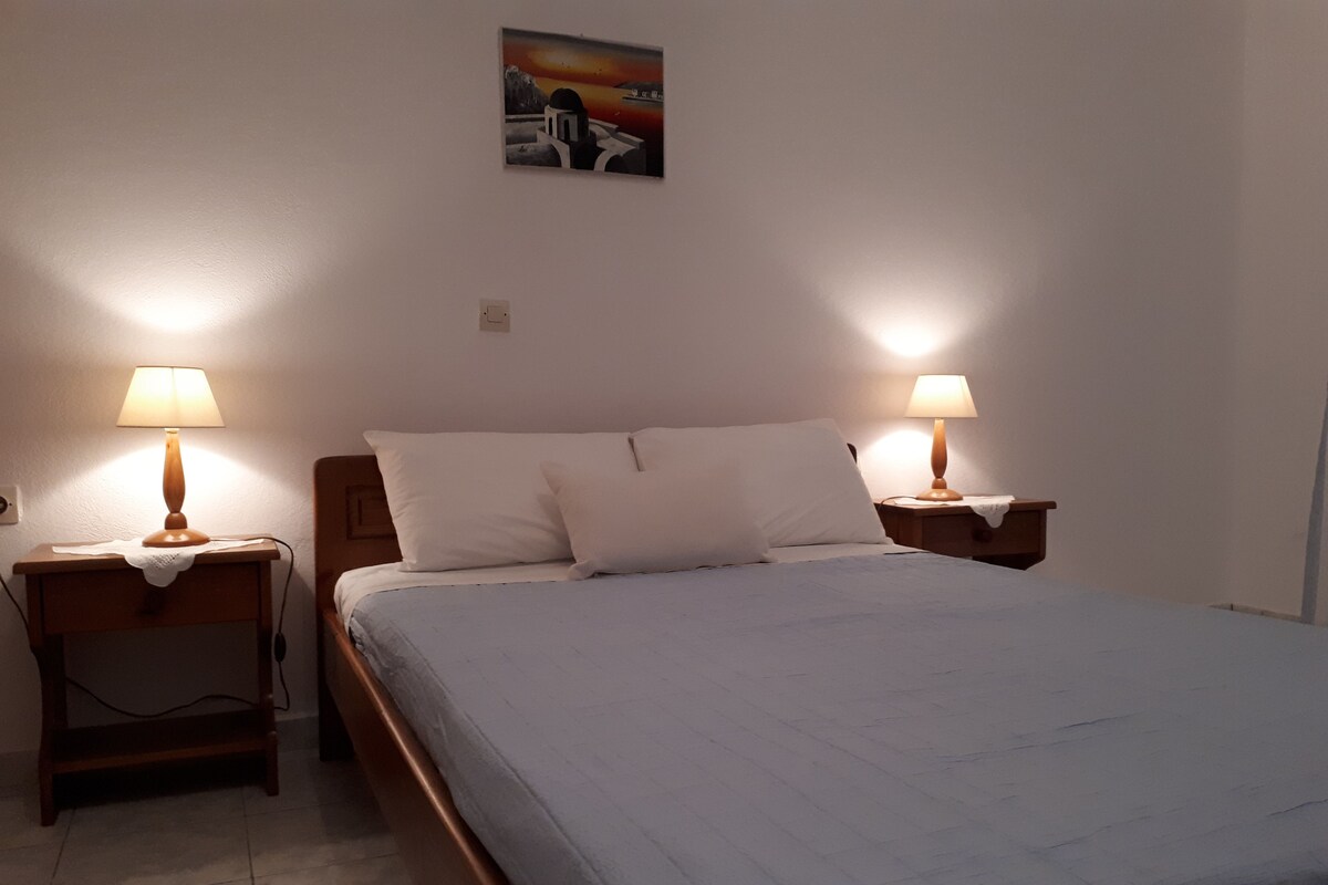 A neatly made bed with two bedside lamps, a small bedside table, and a wall-mounted picture.