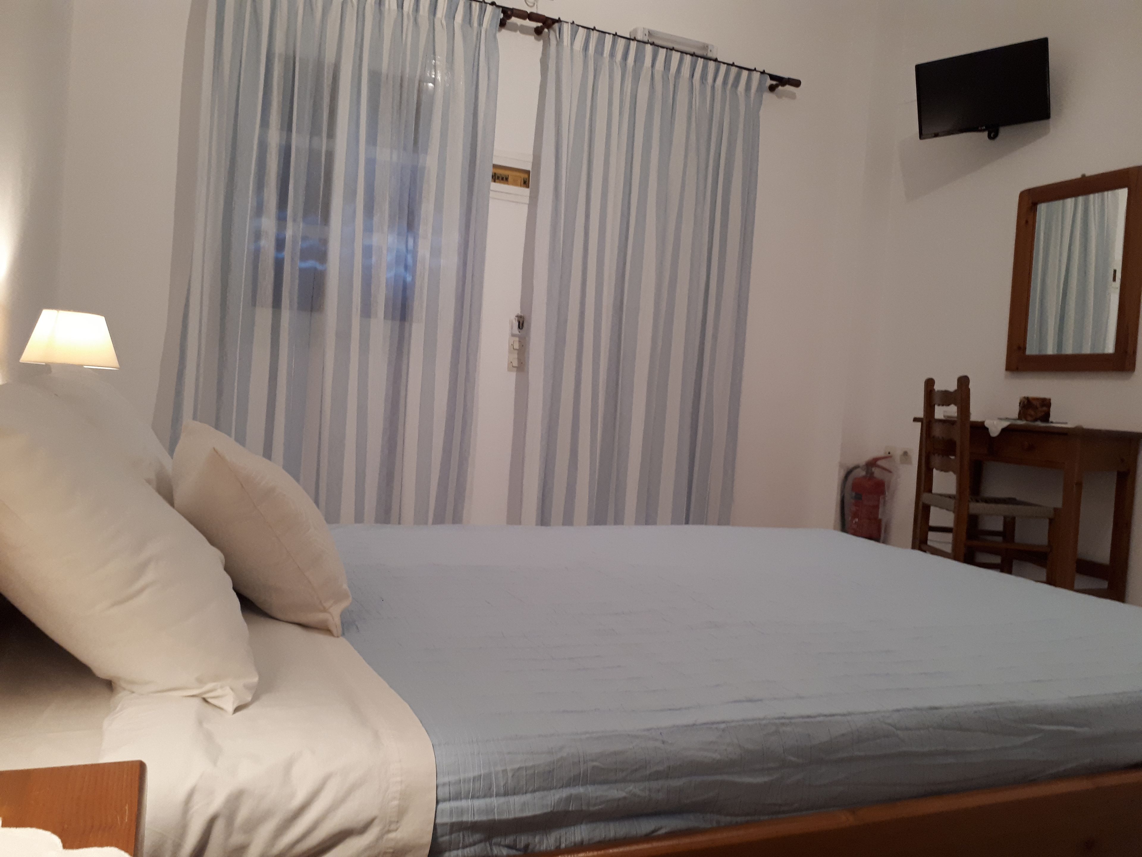 Un dormitorio con cama, mesita de noche, lámpara, silla, escritorio, televisor y ventana con cortinas.