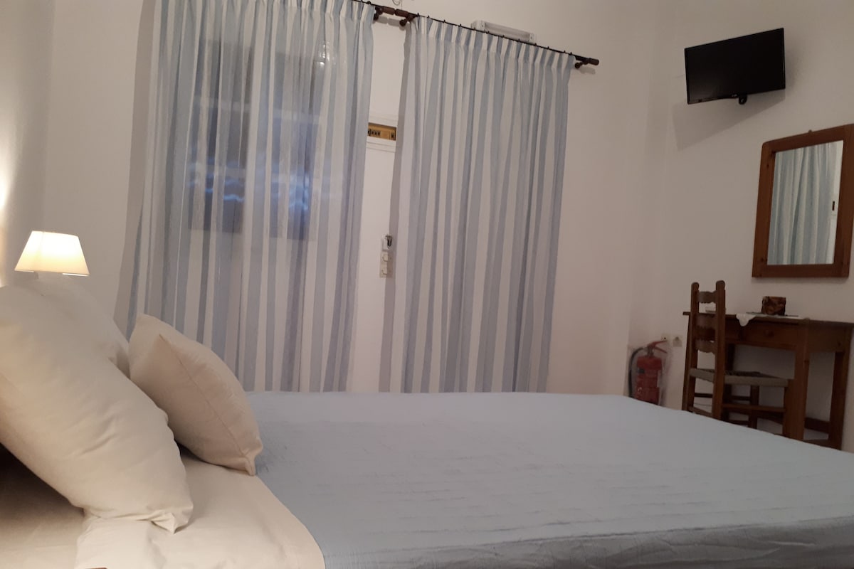 Un dormitorio con cama, mesita de noche, lámpara, silla, escritorio, televisor y ventana con cortinas.