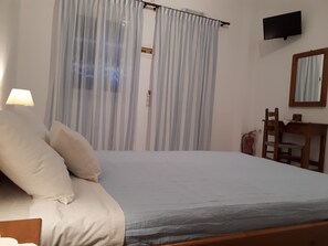 A bedroom with a bed, a nightstand, a lamp, a chair, a desk, a television, and a window with curtains. - Hotel Paradisos Oia (Santorini)