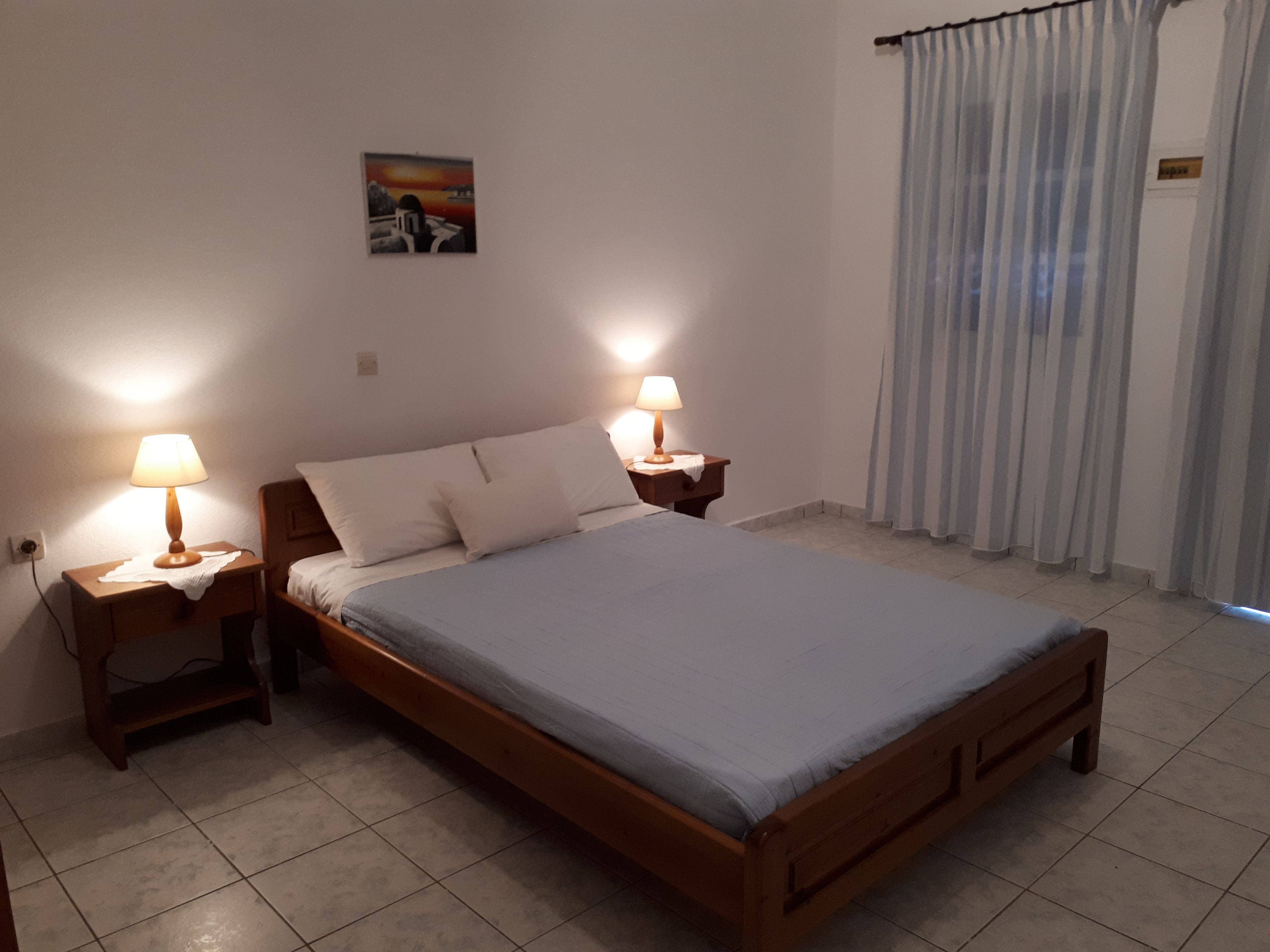 A bedroom with a wooden bed, two bedside tables with lamps, a painting on the wall, and a window with curtains.
