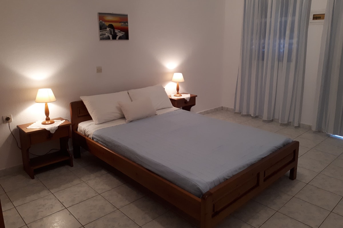 A bedroom with a wooden bed, two bedside tables with lamps, a painting on the wall, and a window with curtains.