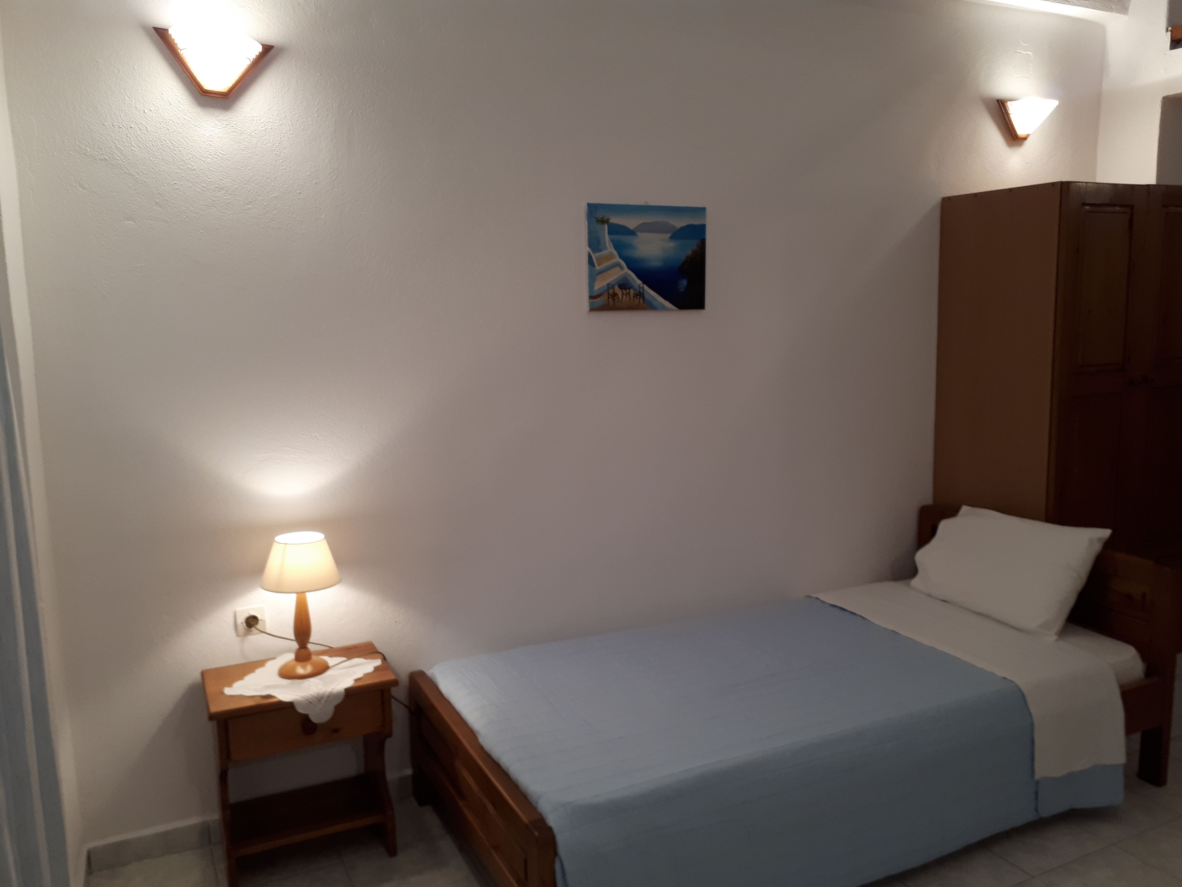 A small bedroom with a single bed, a nightstand with a lamp, a painting on the wall, and a wardrobe.