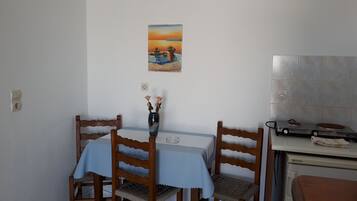 Een eetgedeelte met een tafel voor twee personen, houten stoelen, een vaas met bloemen, een schilderij aan de muur en een kitchenette met een kookplaat.