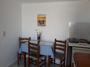 A dining area with a table set for two, wooden chairs, a vase with flowers, a painting on the wall, and a kitchenette with a stove. - Hotel Paradisos Oia (Santorini)