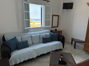 A living room with a sofa, a coffee table, a TV, and a window with an open shutter. - Hotel Paradisos Oia (Santorini)