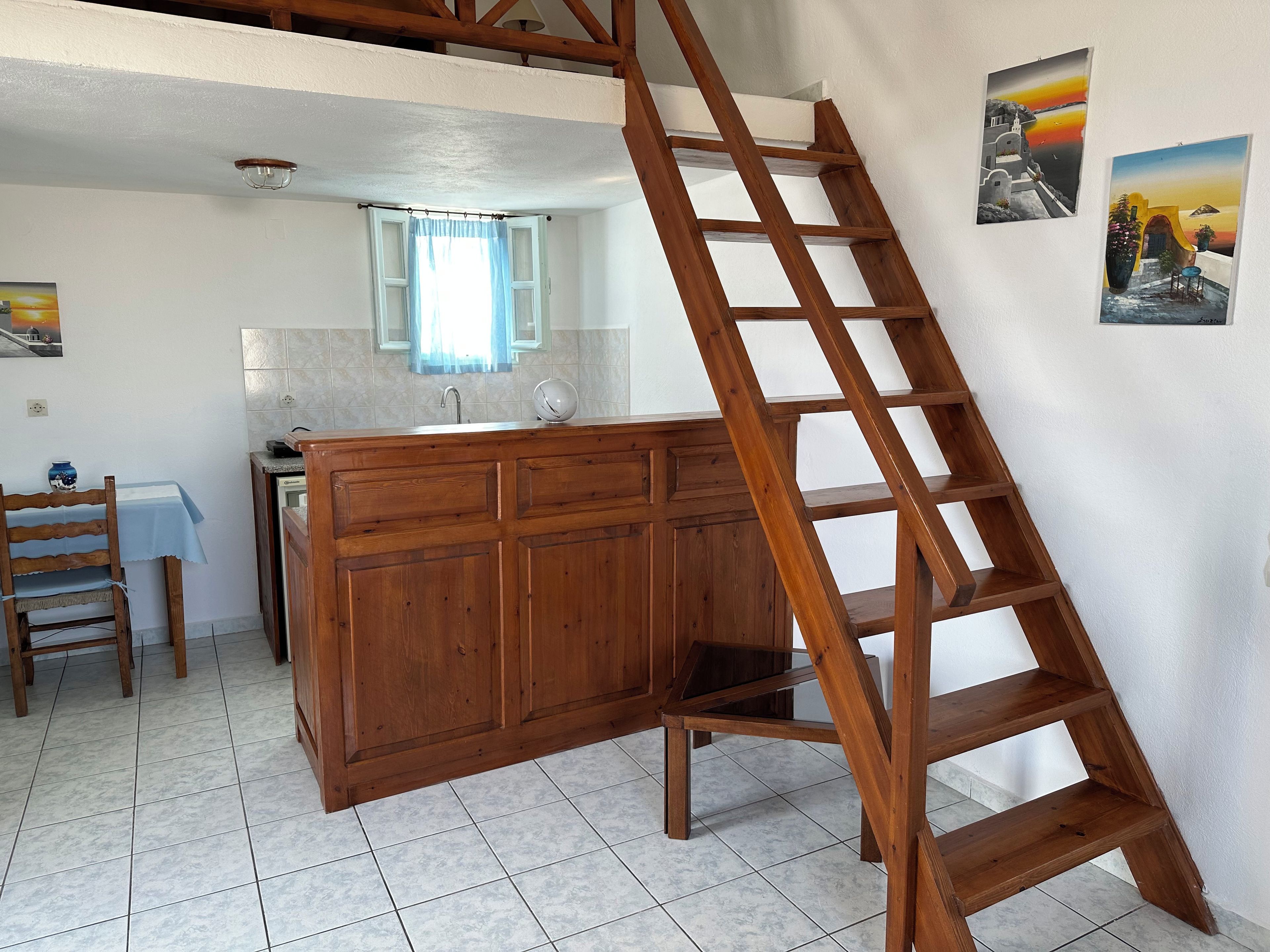 A kitchen with a wooden cabinet, a wooden ladder, a tiled floor, a window with a curtain, and a sink.