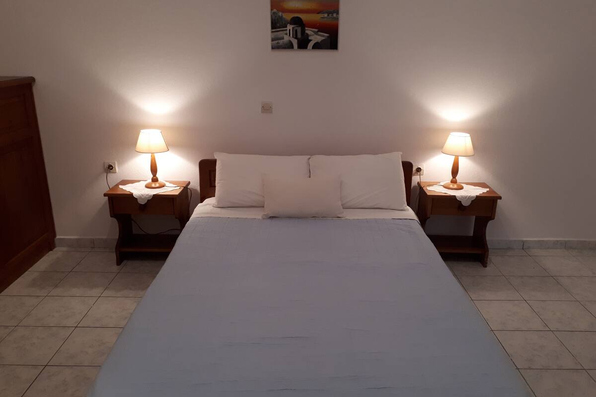A double bed with white linens, two bedside tables with lamps, a small painting on the wall, and a wooden wardrobe.