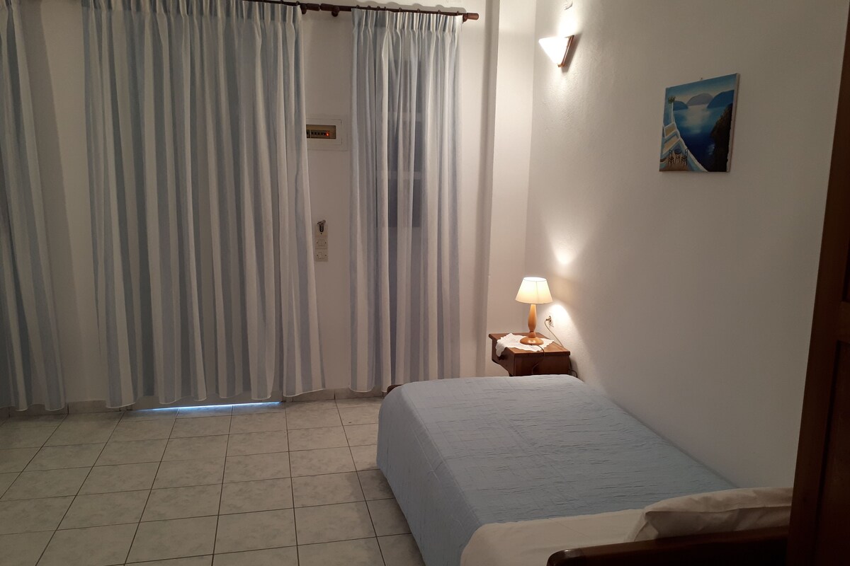 A bedroom with a bed, a nightstand with a lamp, a painting on the wall, and a window with curtains.